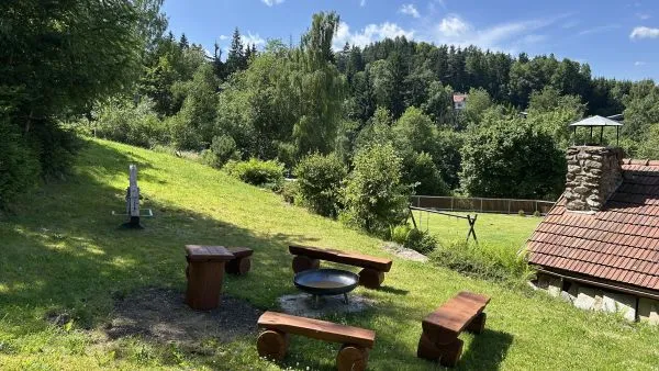 Venkovní ohniště s dřevěným posezením na travnatém svahu nad chalupou Pazderna v létě